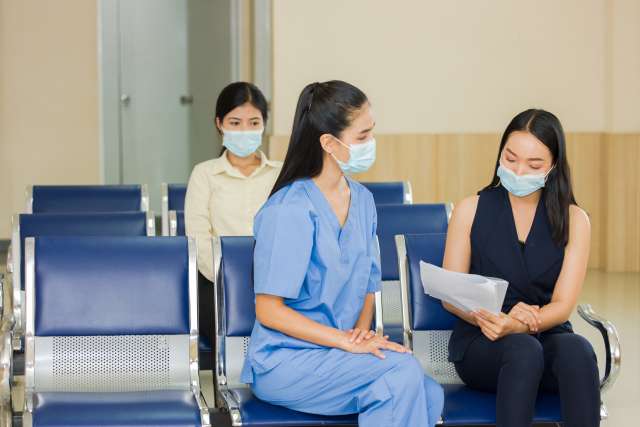 Two women in masks discuss documents while a third waits in a medical setting.