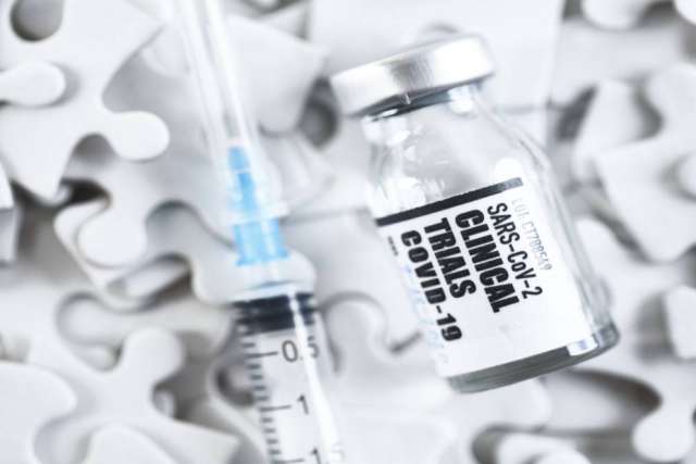  A close-up of a syringe and a glass vial labeled "SARS-CoV-2 CLINICAL TRIALS COVID-19" resting on a background of white puzzle pieces.