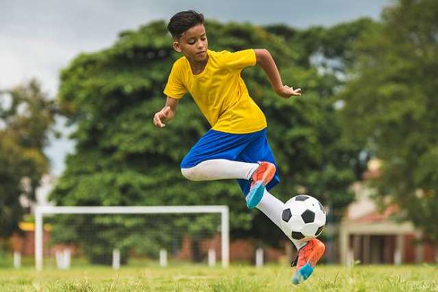 A young boy in a yellow and blue soccer uniform is captured mid-air as he runs and kicks a black and white soccer ball on a grassy field.