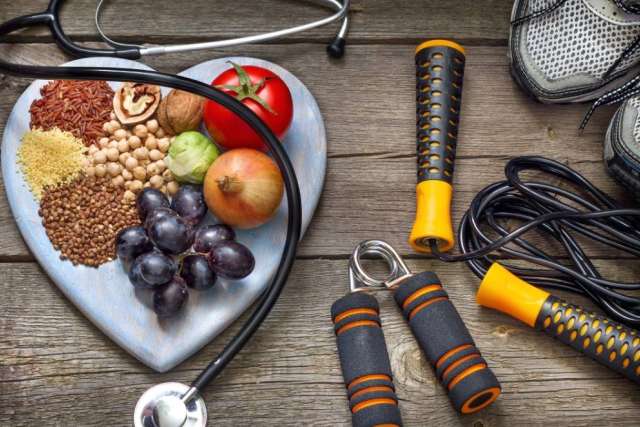 Heart-shaped tray with grains, nuts, fruits, and vegetables next to a stethoscope, jump rope, and sneakers on a wooden surface.