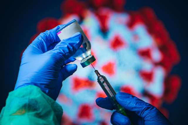 Gloved hands drawing a COVID-19 vaccine dose from a vial with a syringe, with a virus illustration in the background.