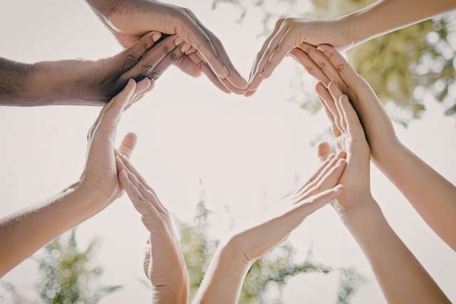 Multiple hands forming a heart shape together against the bright sky.