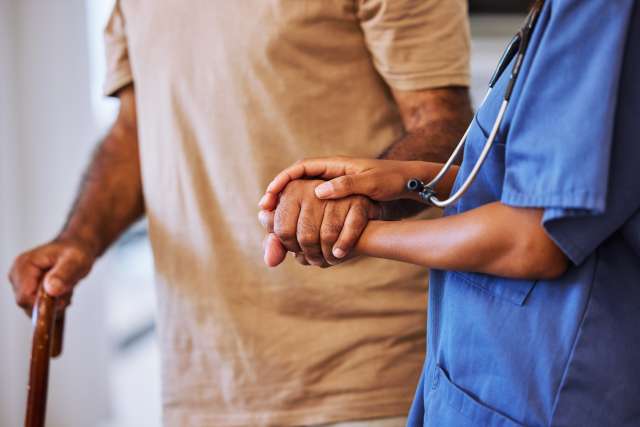 A caregiver holds a patient's hand, helping the patient to walk.