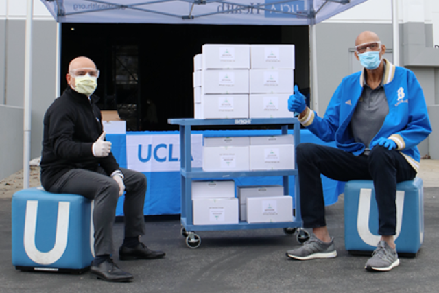 Two men sitting on UCLA branded stools under a tent with boxes in front of them.