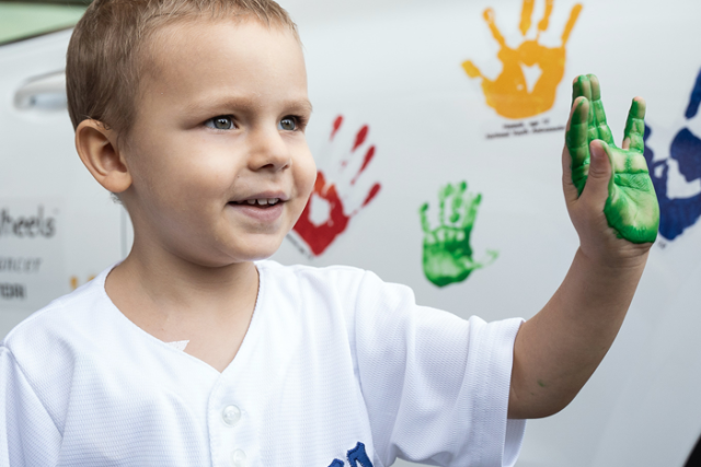 A young boy in a "Dodgers" shirt, smiling, shows his green-painted hand while standing next to a white car covered in colorful handprints.