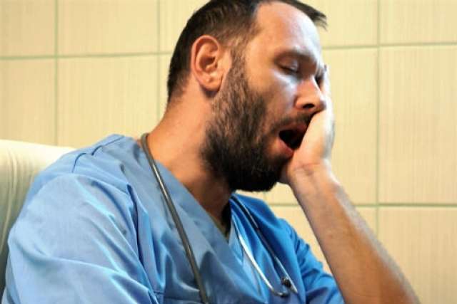 A male medical professional in blue scrubs, with a beard and stethoscope, yawns with his eyes closed and hand on his face.