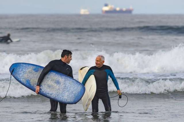 Two men wearing wetsuits while carrying surfboards and walking through shallow water on the beach