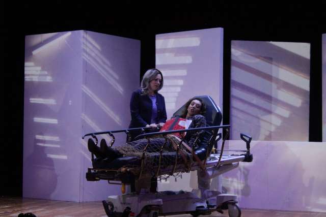 An image of a woman on stage standing next to another woman who's restrained in a hospital bed