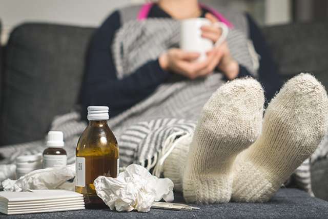 A person is resting on a sofa under a blanket, wearing white wool socks, with medicine bottles, tissues, and a thermometer visible in the foreground.