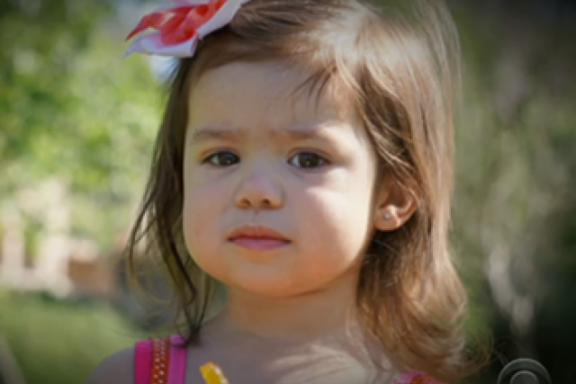  A headshot of a young girl with brown hair and a red and white bow in her hair. She is looking directly at the camera with a neutral expression.