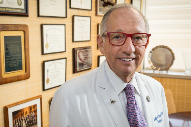 Ronald W. Busuttil, MD, PhD standing in front of a yellow wall with many picture frames, smiling and looking at the camera