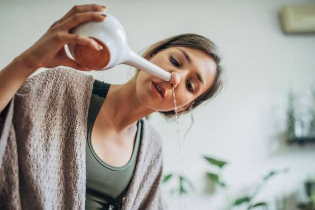 A woman uses a ceramic neti pot to perform nasal irrigation, tilting her head to allow water to flow from one nostril to the other.