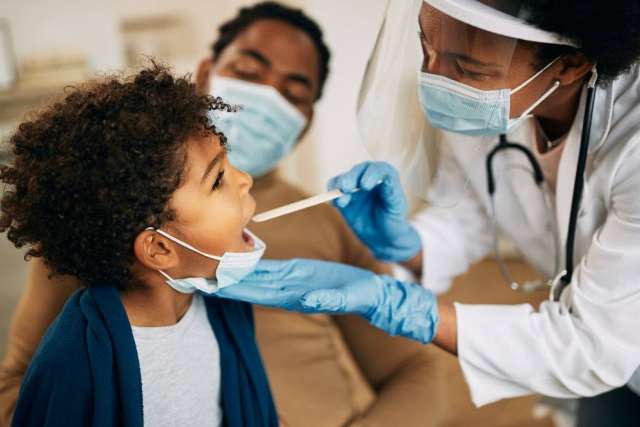 Healthcare professional wearing gloves and a face shield performing a throat swab test on a child, with another person in the background.