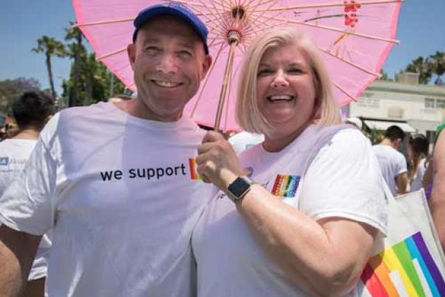 A smiling man and woman stand close under a pink umbrella, both wearing white "we support" shirts with a rainbow graphic, likely at a parade.
