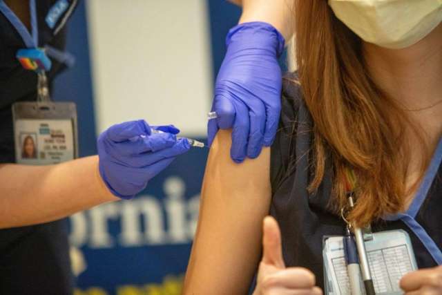 Healthcare worker in gloves gives a COVID-19 vaccine shot to a masked person giving a thumbs-up
