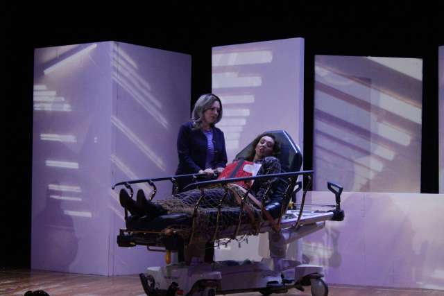 An image of a woman on stage standing next to another woman who's restrained in a hospital bed