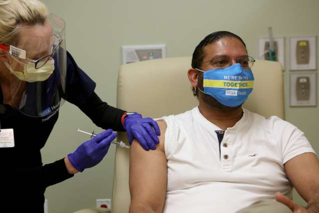 A doctor wearing a mask and gloves gives Dr. Robert Cherry a vaccination in his arm. He is wearing a face mask and a white polo shirt.