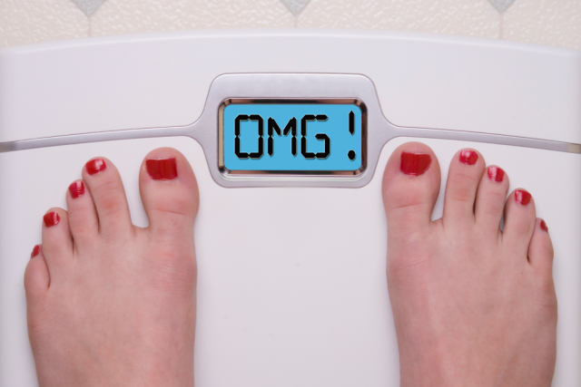 A pair of feet with painted red toenails stands on a white scale. The digital display reads "OMG!"