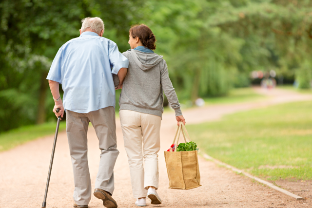 Caregiver assisting an elderly man with a cane as they walk outdoors on a park path, symbolizing support and independence.
