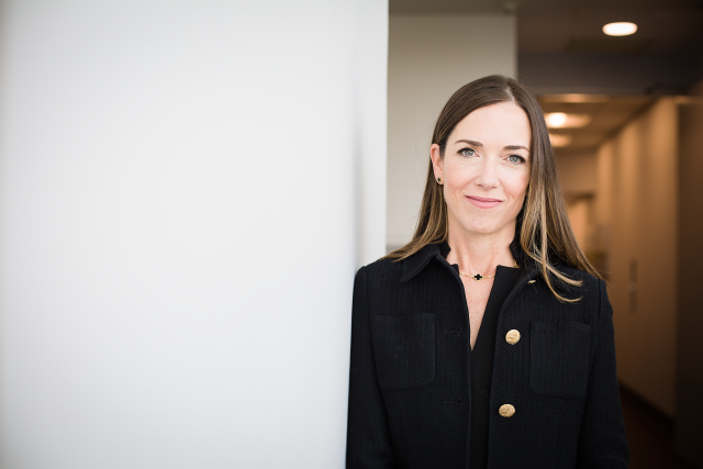 Dr. Sara Hurvitz leaning on a white wall, wearing a black dress and a black coat with golden buttons over it.