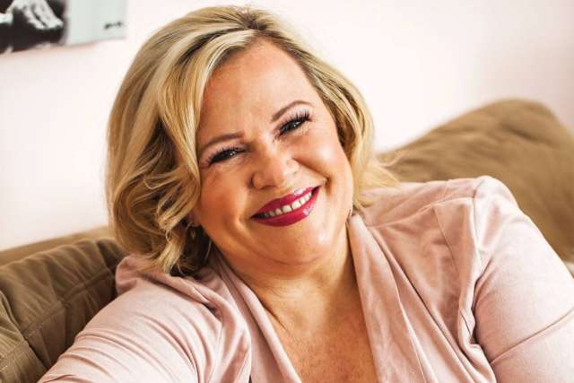 A person with blonde hair sits on a couch wearing a light pink top in a relaxed, indoor setting.