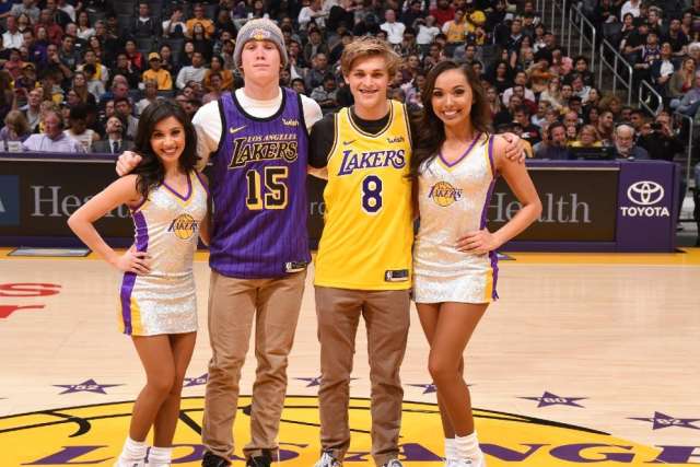 Two people wearing Los Angeles Lakers jerseys posing with two Lakers cheerleaders on the basketball court during a game.