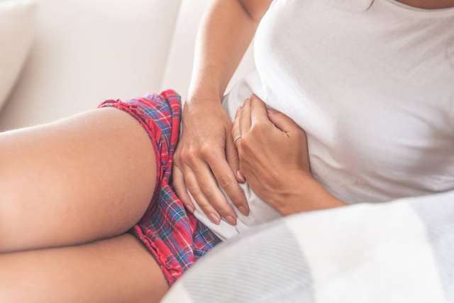 Woman in a white shirt and plaid shorts holding her stomach, looking uncomfortable.