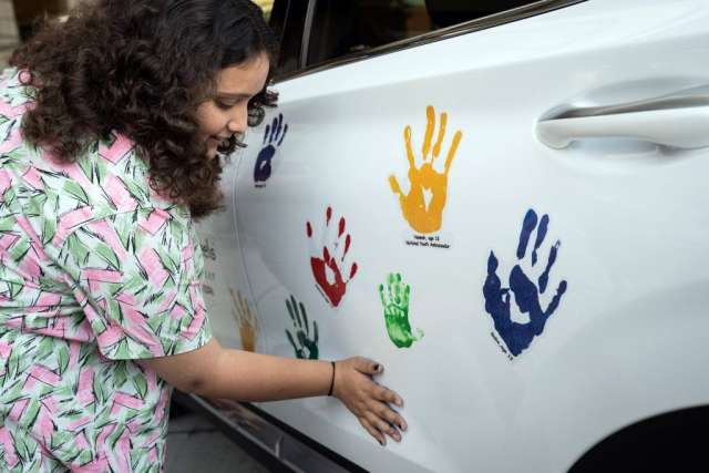 A young woman in a floral shirt smiles while gently touching one of several colorful handprints on the white side of a car.