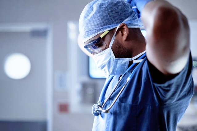 Surgeon wearing scrubs, mask, and gloves prepares for procedure in a hospital operating room.