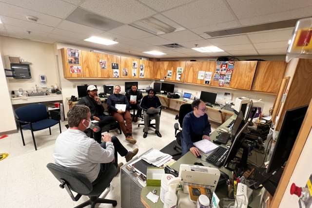 Small office with several people working at computer stations around a central desk.