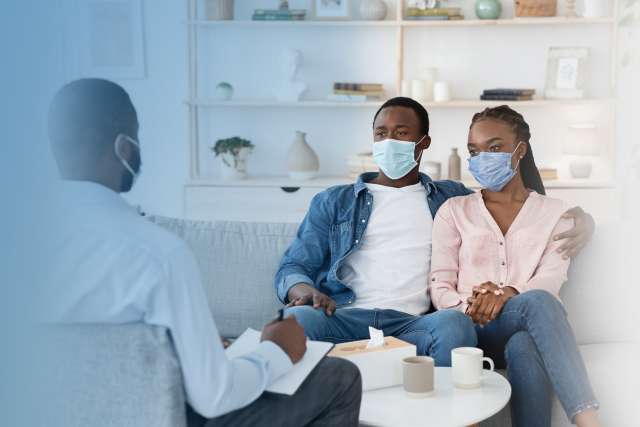 Family Therapist Talking To Black Couple Sitting In Protective Masks On Couch At Office