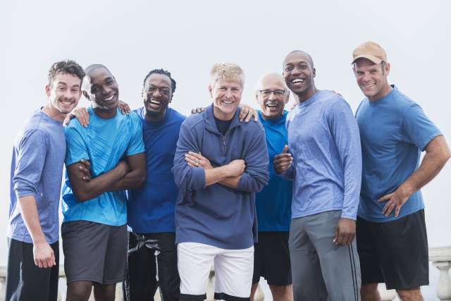 Diverse group of men wearing casual blue shirts, standing together outdoors. Mixed ages, 20s to 70s. They are confident and happy, smiling at the camera.