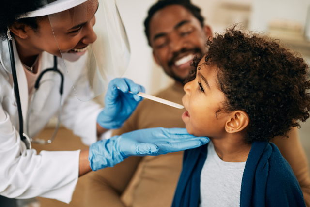 African American doctor with face mask examining boy's throat during a home visit.