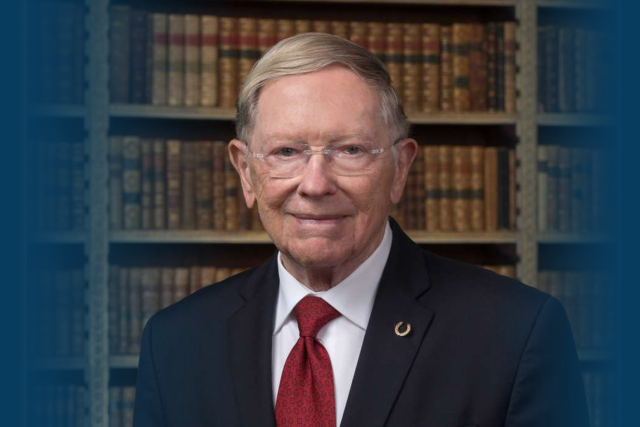 Older man in dark suit and red tie smiling in front of bookshelves.
