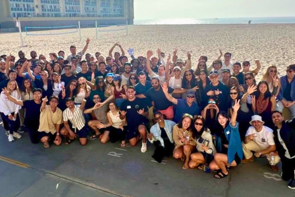 A large group of DAPM residents pose on a beach. Many are smiling and raising their hands. The background shows the ocean and a building.
