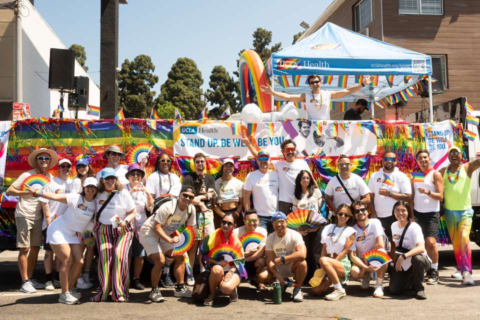 A large group photo in front of the UCLA Health float, with many holding Pride flags and rainbow fans