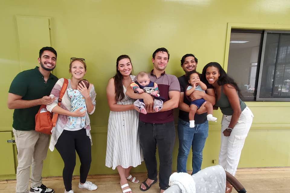 Seven smiling people, including two holding babies, stand together in front of a green wall indoors.