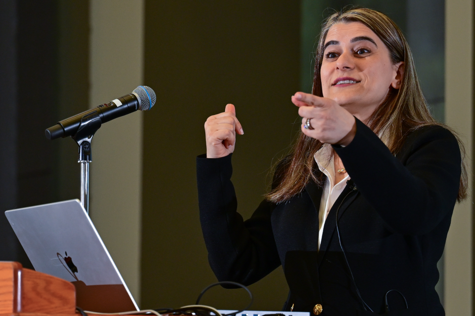 Person speaking at podium, gesturing with hands, microphone and laptop on lectern.