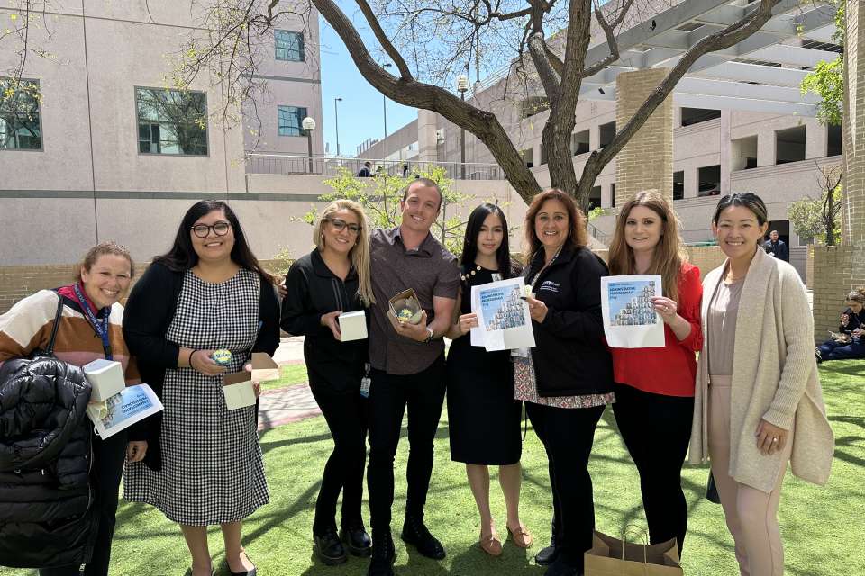 A group of smiling administrative professionals pose for a group photo outdoors, holding up papers. The background includes a tree and a building.