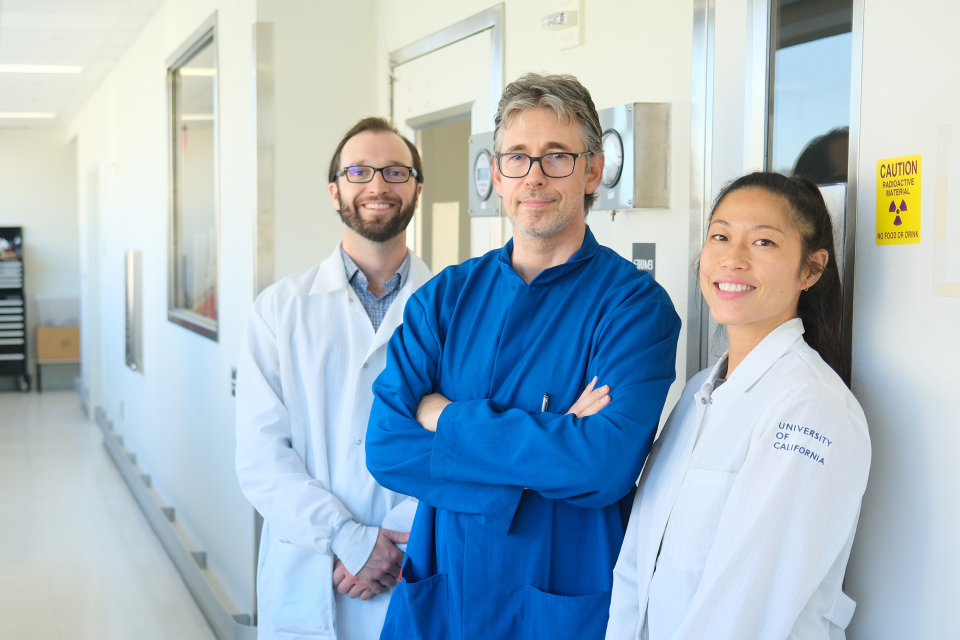 Two male doctors and one female doctor smiling in a hospital hallway wearing the lab coats, two wearing white and the middle doctor wearing blue.