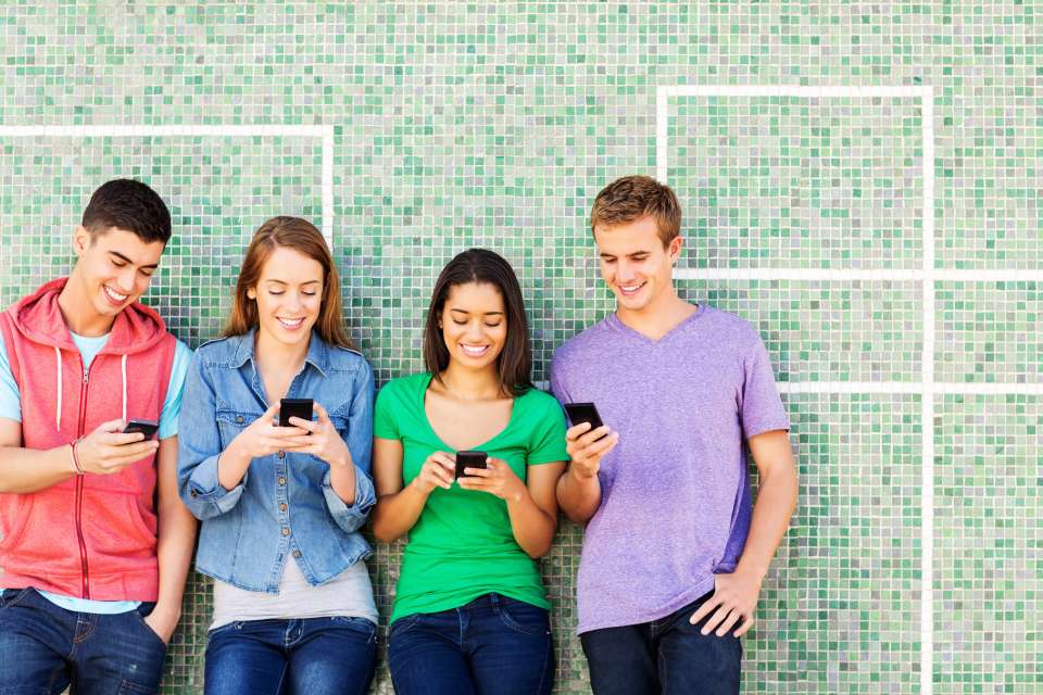 Teens against a wall looking at phones: four teens standing and using phones