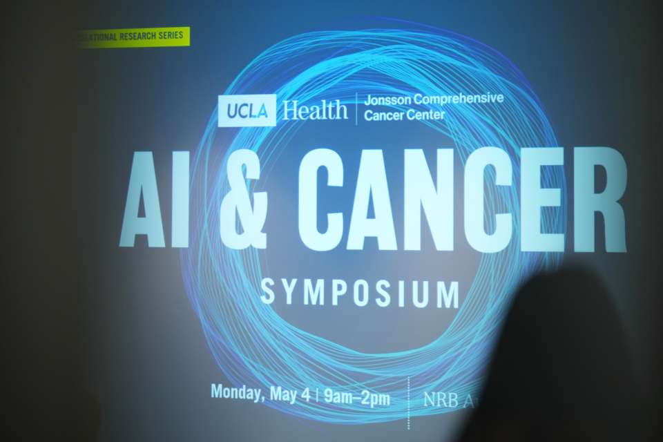 Silhouettes of cancer researchers standing in front of a digital sign that says "Ai & Cancer Symposium"