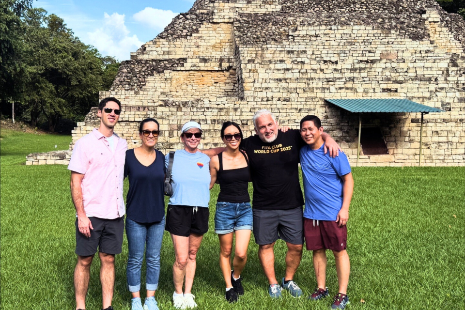 Group of six people posing in front of a stone pyramid in a grassy area.