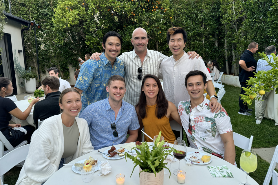 A group of seven Orthopaedic surgery residents are gathered around a table outdoors, with a garden setting in the background.