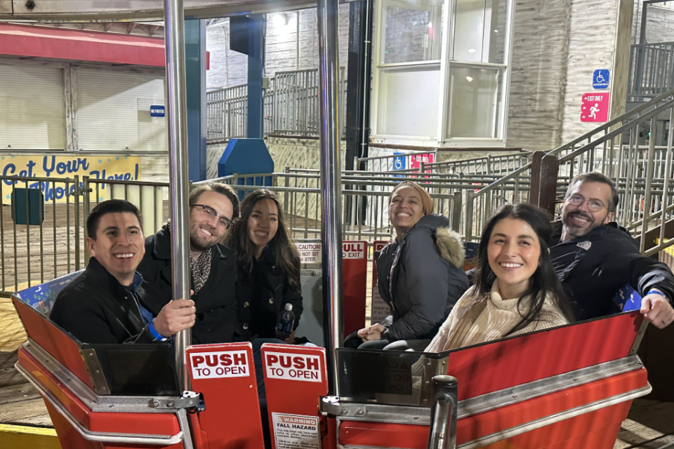 Six smiling people sit in red carnival ride seats at night, bundled in coats. 