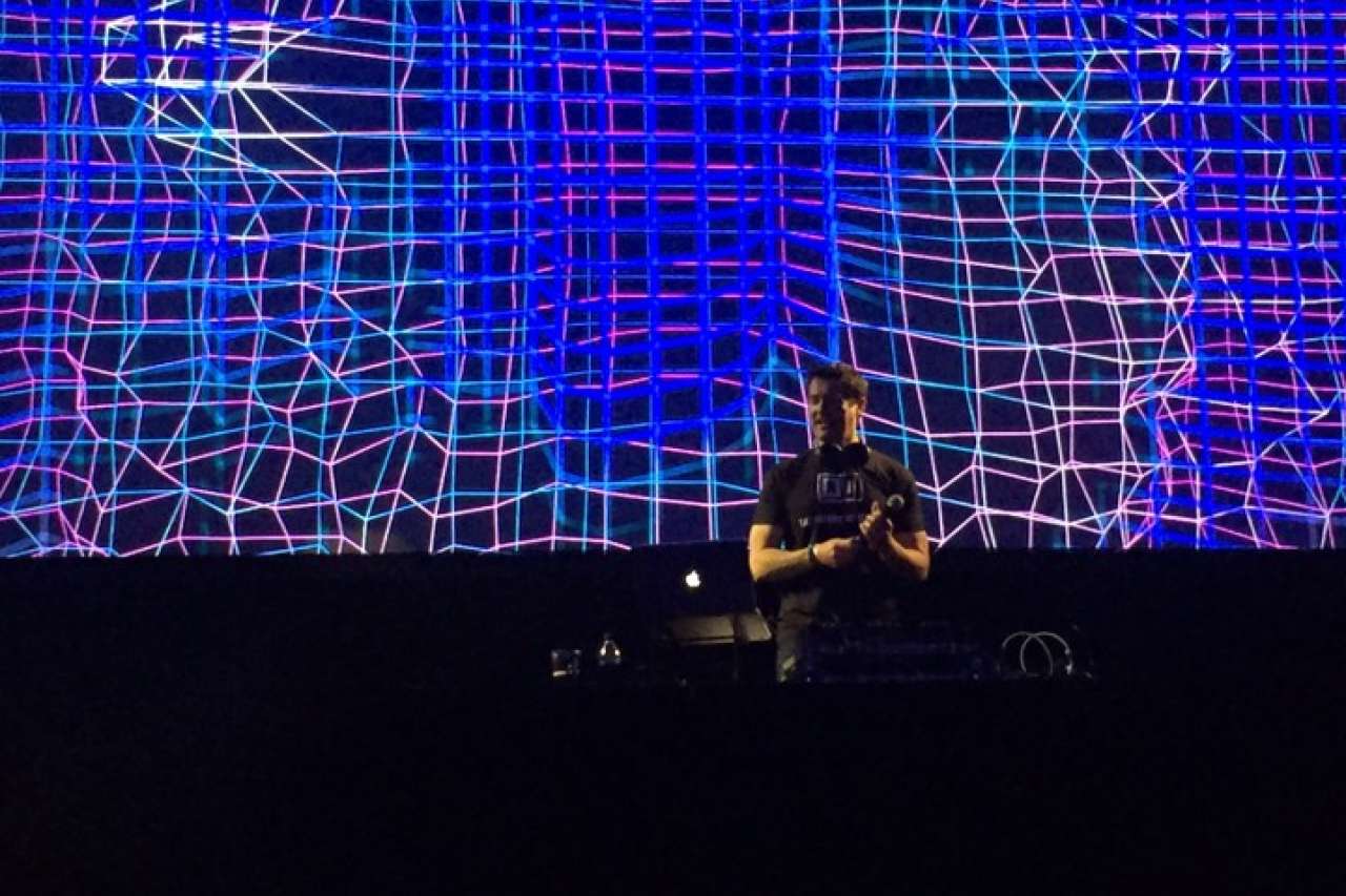 Dr. Tony Aldave wears a black T-shirt and holds a microphone on a vibrant DJ stage.