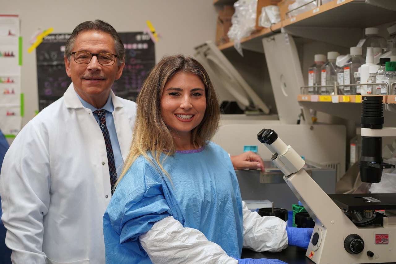 scientist Donald Kohn on the left wearing white lab coat and scientist Eva Segura on the right wearing a lab protective gear in a lab next to a microscope