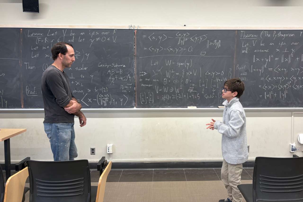 A boy and a teacher talk in front of a blackboard covered with mathematical equations.