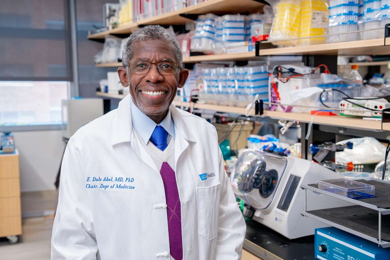 Doctor Abel in a white lab coat standing in a laboratory with equipment and shelves in the background