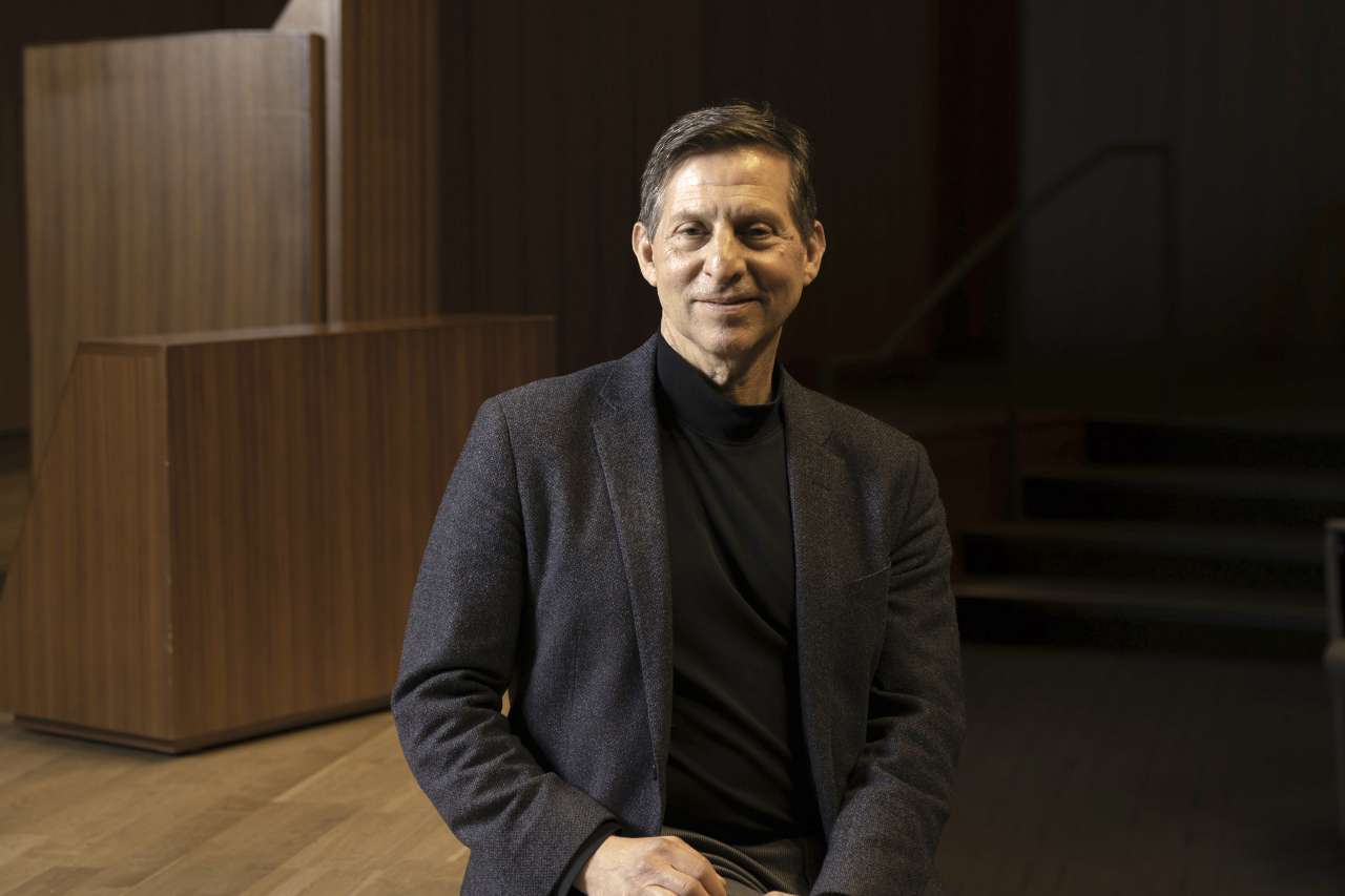 Dr. Paul Mischel seated and wearing a blazer on stage, hands clasped and slight smile